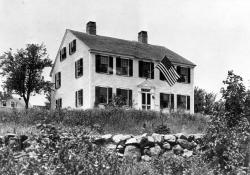 Historic white house with black shutters and an American flag, surrounded by grass and a stone wall.