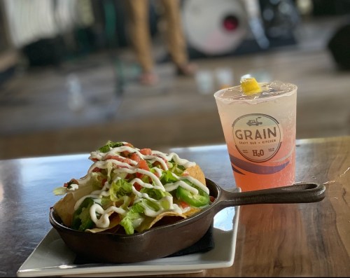 A skillet of nachos topped with lettuce, tomatoes, and sour cream, alongside a refreshing drink with a lemon slice.