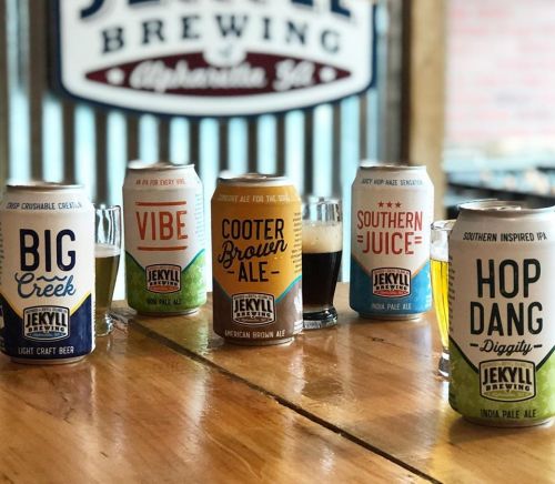 A selection of Jekyll Brewing beers on a wooden table, featuring various colorful cans and small tasting glasses.