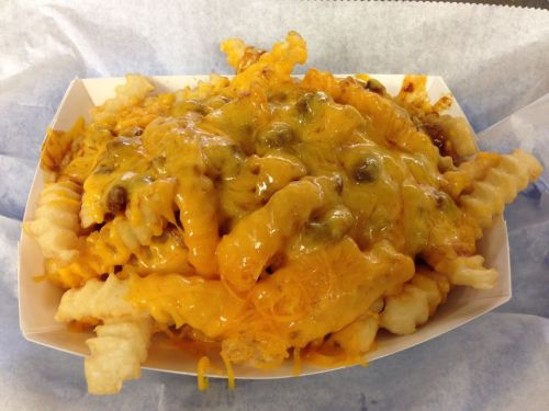 A serving of crinkle-cut fries topped with melted cheese and green toppings in a paper tray.