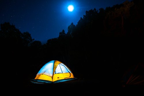 A brightly lit tent glows under a full moon in a dark, starry night sky surrounded by trees.