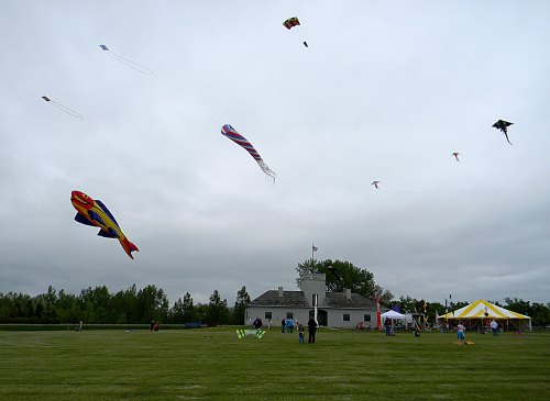 Colorful kites flying in a cloudy sky over a grassy field, with people and a tent in the background.