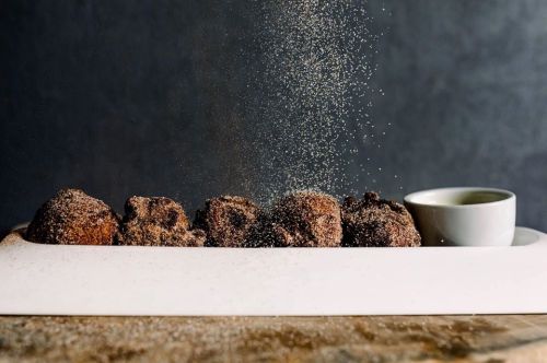 A plate of chocolate-dusted doughnuts with a small cup of sauce, sugar being sprinkled over them.