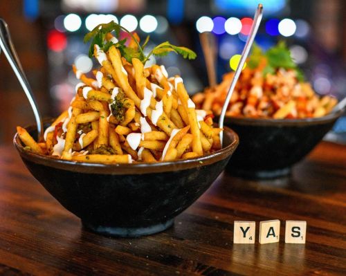 Two bowls of seasoned fries topped with sauces and garnishes, with letter tiles spelling "Y.A.S." in the foreground.