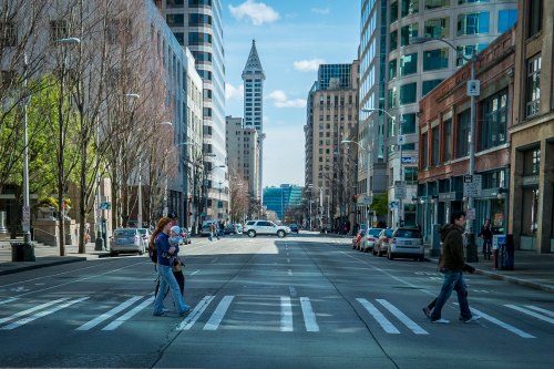 A city street with pedestrians crossing, tall buildings lining the road, and a distant view of a tower.