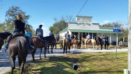 A group of people on horseback gather outside a general store on a sunny day.