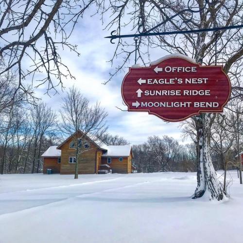 A wooden sign with directional arrows points to various locations near a snow-covered cabin and bare trees.