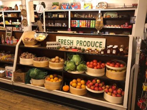 A display of fresh produce in baskets, featuring apples, oranges, potatoes, and cabbages in a vintage store setting.