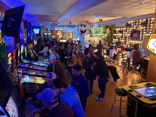 A lively arcade filled with people playing pinball and arcade games, colorful lights illuminating the space.