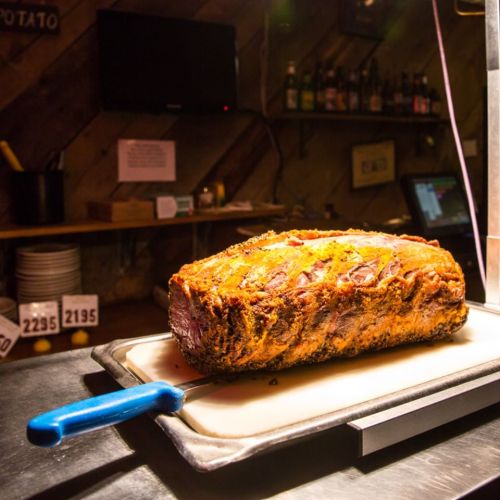 A large, seasoned roast resting on a cutting board with a blue-handled knife beside it, in a rustic setting.