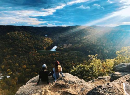 Two people sit on a rocky ledge, overlooking a valley with a river and colorful autumn trees under a dramatic sky.