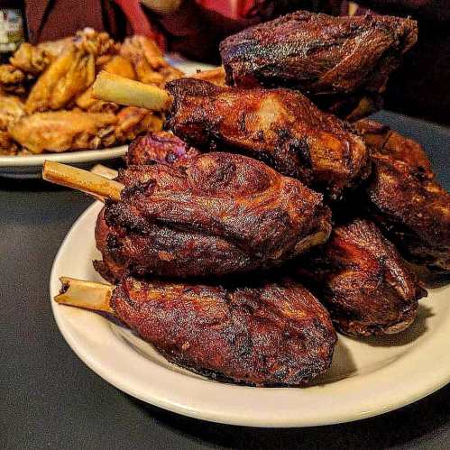 A plate stacked high with crispy, smoked meat on bone, with a side of chicken wings in the background.