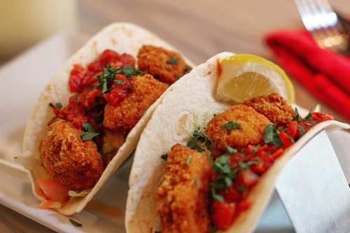 Two crispy fish tacos topped with fresh salsa, cilantro, and a wedge of lemon on a white plate.
