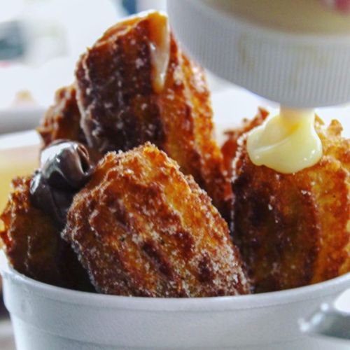 A close-up of crispy, golden churros in a cup, drizzled with chocolate and cream sauces.