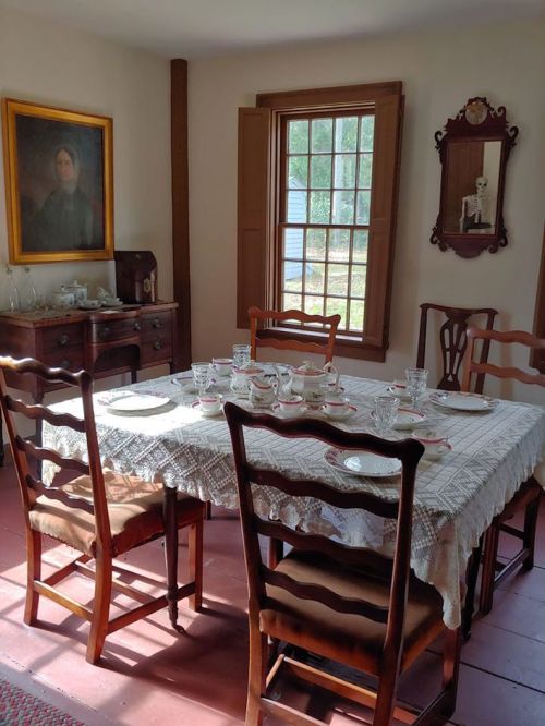 A cozy dining room with a lace tablecloth, set for tea, featuring wooden chairs and a portrait on the wall.