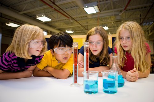 Four children in safety goggles observe colorful liquids in beakers during a science experiment.