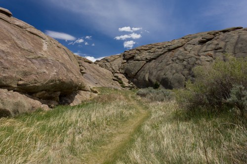 A narrow path winds through rocky terrain and tall grass under a blue sky with scattered clouds.