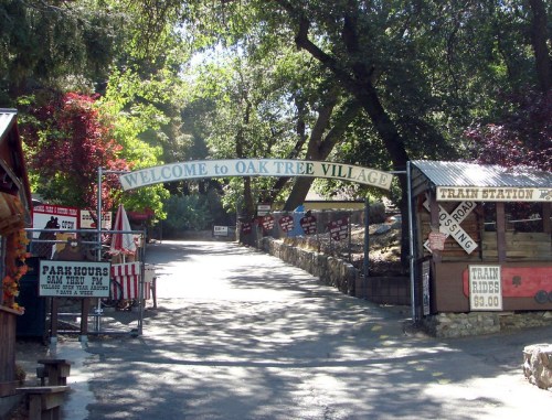 Entrance to Oak Tree Village with a welcoming sign, surrounded by trees and rustic buildings. Train station visible ahead.