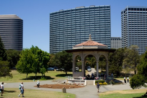 A park scene featuring a gazebo surrounded by greenery and modern buildings in the background on a sunny day.