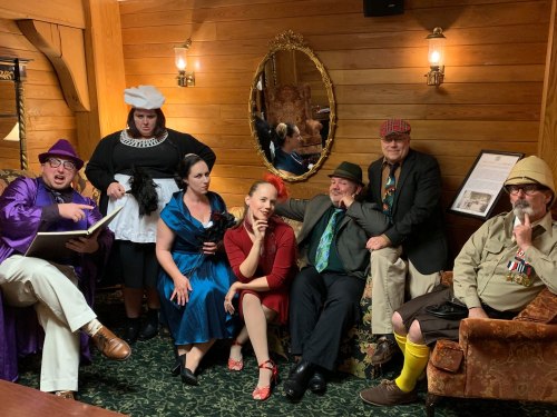 A group of people in vintage costumes poses in a cozy, wooden room with a mirror and decorative furnishings.