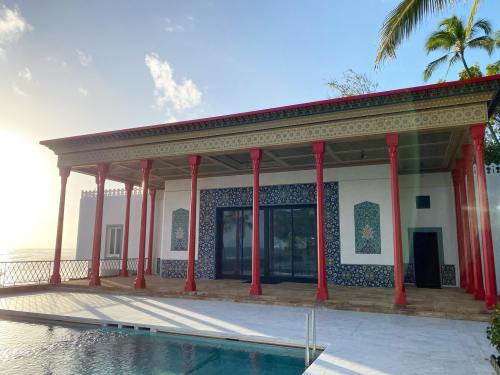 A modern building with red columns and intricate tile work, overlooking a pool and the ocean at sunset.