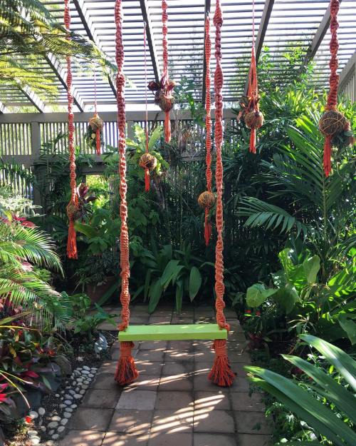 A vibrant garden with a macramé swing hanging from a pergola, surrounded by lush green plants and colorful foliage.
