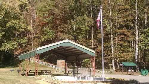 A covered picnic area with a flagpole, surrounded by trees and a gravel path in a serene outdoor setting.
