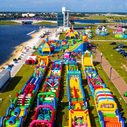 Aerial view of a colorful inflatable water park with slides and obstacles by a lake, bustling with visitors.