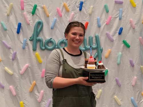 A smiling woman holds a dessert-themed trophy in front of a colorful, candy-themed backdrop.
