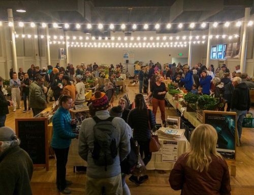 A bustling indoor market with people browsing tables filled with fresh produce and goods, illuminated by string lights.