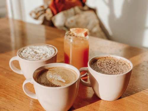 Three cups of coffee and a jar of orange drink on a wooden table, with sunlight casting soft shadows.