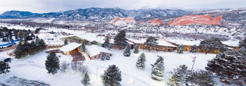 A snowy landscape featuring a lodge surrounded by trees and mountains, with red rock formations in the background.