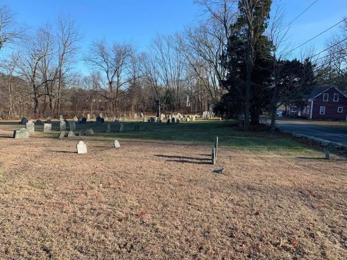 A grassy cemetery with numerous gravestones, surrounded by bare trees and a road in the background.