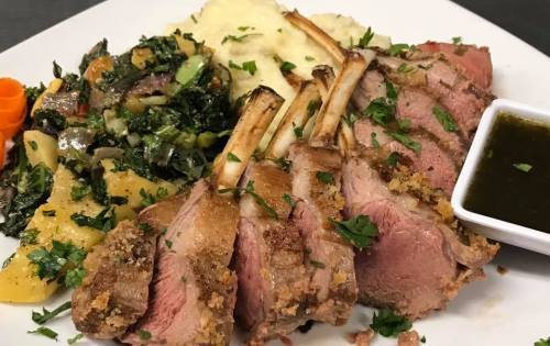 A plate of sliced lamb chops with mashed potatoes, sautéed greens, and a small dish of sauce on the side.