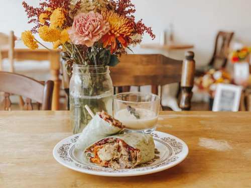 A plate with a green wrap filled with ingredients, a glass of dessert, and a vase of flowers on a wooden table.
