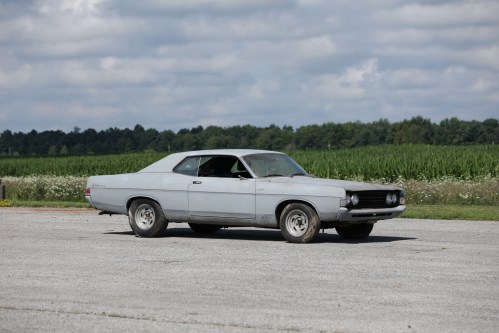 A vintage gray car parked on a gravel surface with a green field in the background under a cloudy sky.