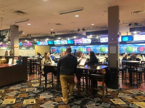 A lively bowling alley with people socializing at tables and bowling lanes in the background, illuminated screens above.