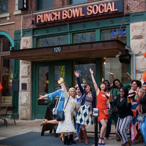 A group of women joyfully posing in front of the Punch Bowl Social entrance, waving and smiling.