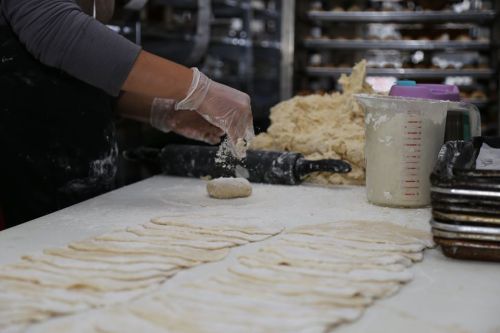 A person in gloves rolls dough on a floured surface, with cut dough pieces and baking trays in the background.