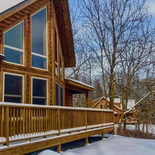 A log cabin with large windows, surrounded by snow-covered trees and a wooden deck, under a clear blue sky.