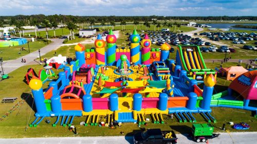 A colorful inflatable bounce park with various slides and structures, set in a grassy area with cars parked nearby.