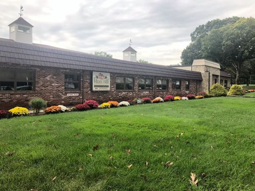 A brick building with a sign, surrounded by colorful flowers and green grass under a cloudy sky.