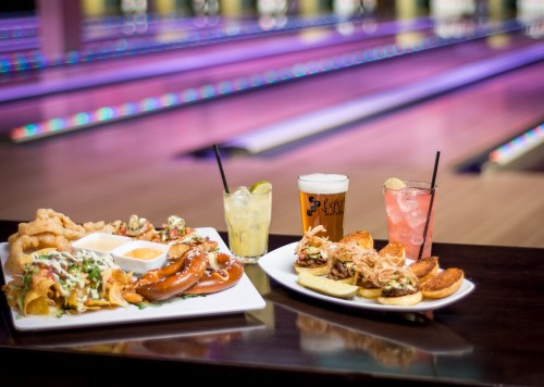 A table with nachos, sausages, sliders, and drinks, set against a colorful bowling alley backdrop.