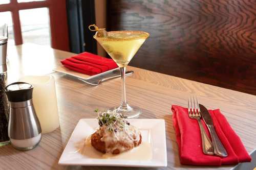 A martini with olives beside a plate of creamy dish garnished with microgreens, set on a wooden table with red napkins.
