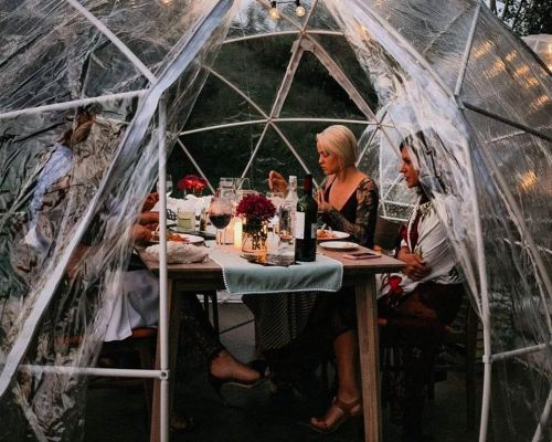 A group of people dining inside a clear dome, surrounded by flowers and warm lighting, enjoying a cozy meal together.