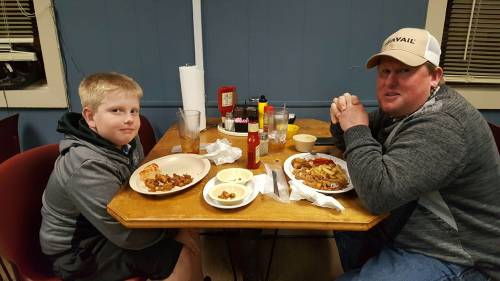 A man and a boy sit at a table with plates of food, drinks, and condiments in a casual dining setting.
