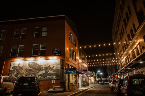 Nighttime view of a street with string lights, featuring the Harbor Inn and a mural of fish on a building.