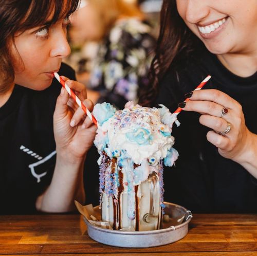 Two women share a colorful, overflowing milkshake topped with whipped cream and sprinkles, each holding a straw.