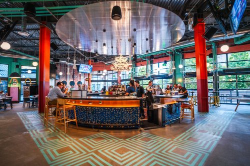 A vibrant bar area with colorful decor, patrons seated at the bar, and large windows letting in natural light.