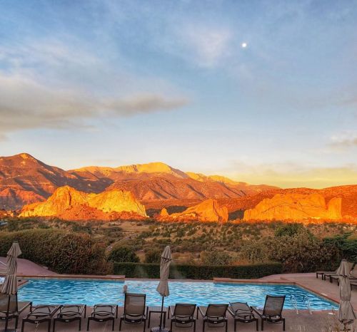 Sunset over mountains with a pool in the foreground, surrounded by lounge chairs and lush greenery.